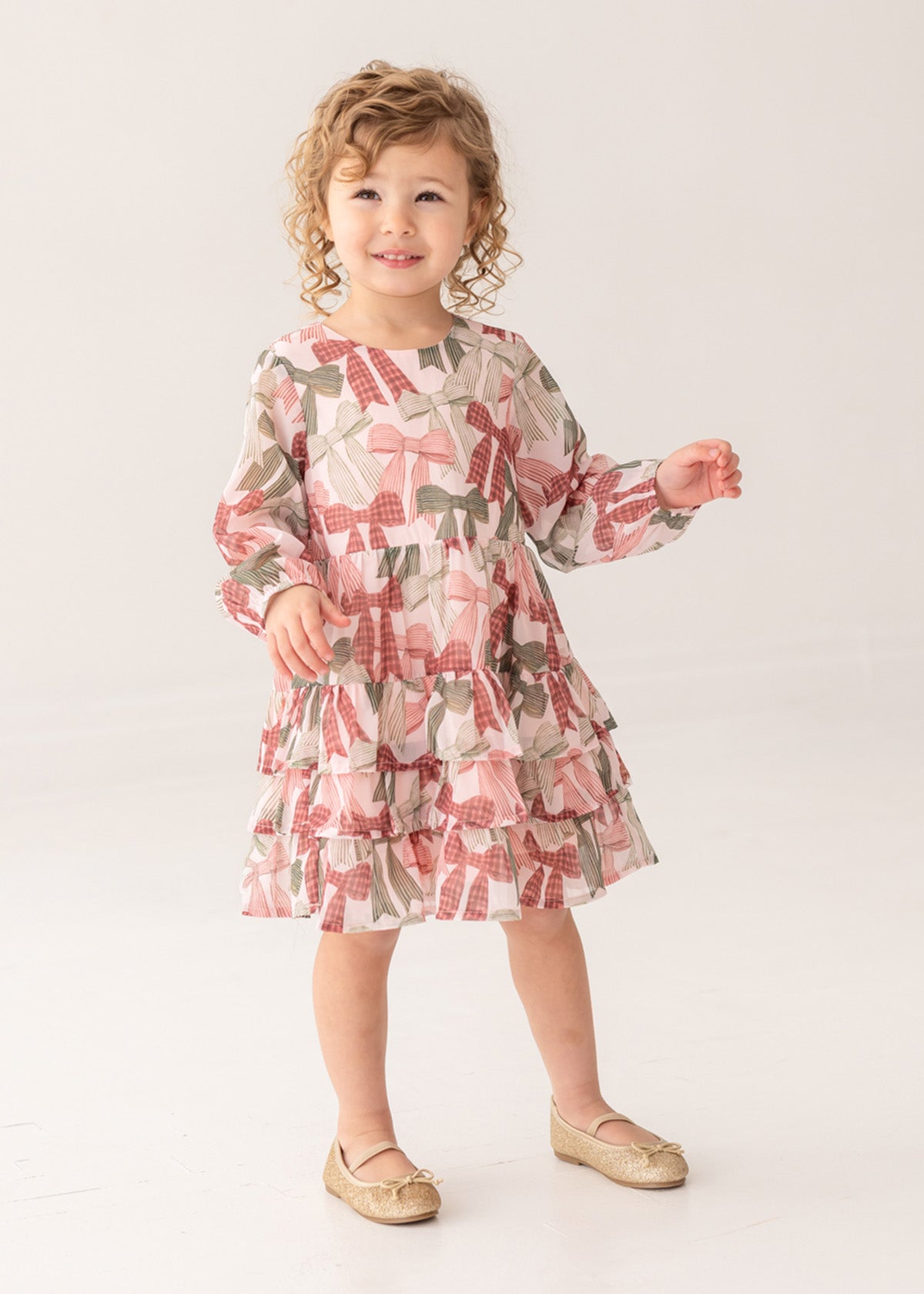 A young child with curly hair wears the Mabel and Honey Bow-tiful Dress, featuring a pink and green geometric pattern, paired with gold shoes, standing and smiling against a plain light background.