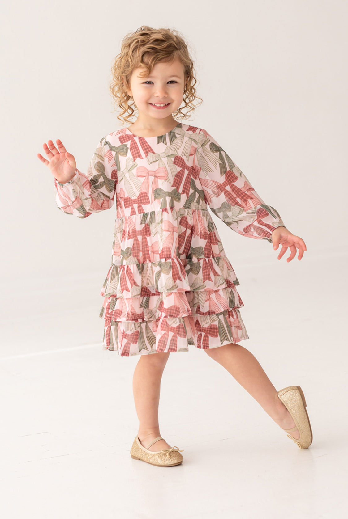 A curly-haired girl, smiling and waving, poses on one foot in a bright studio wearing the Bow-tiful Dress by Mabel and Honey with pink and olive tiered ruffles, paired with gold flats for a charming look.