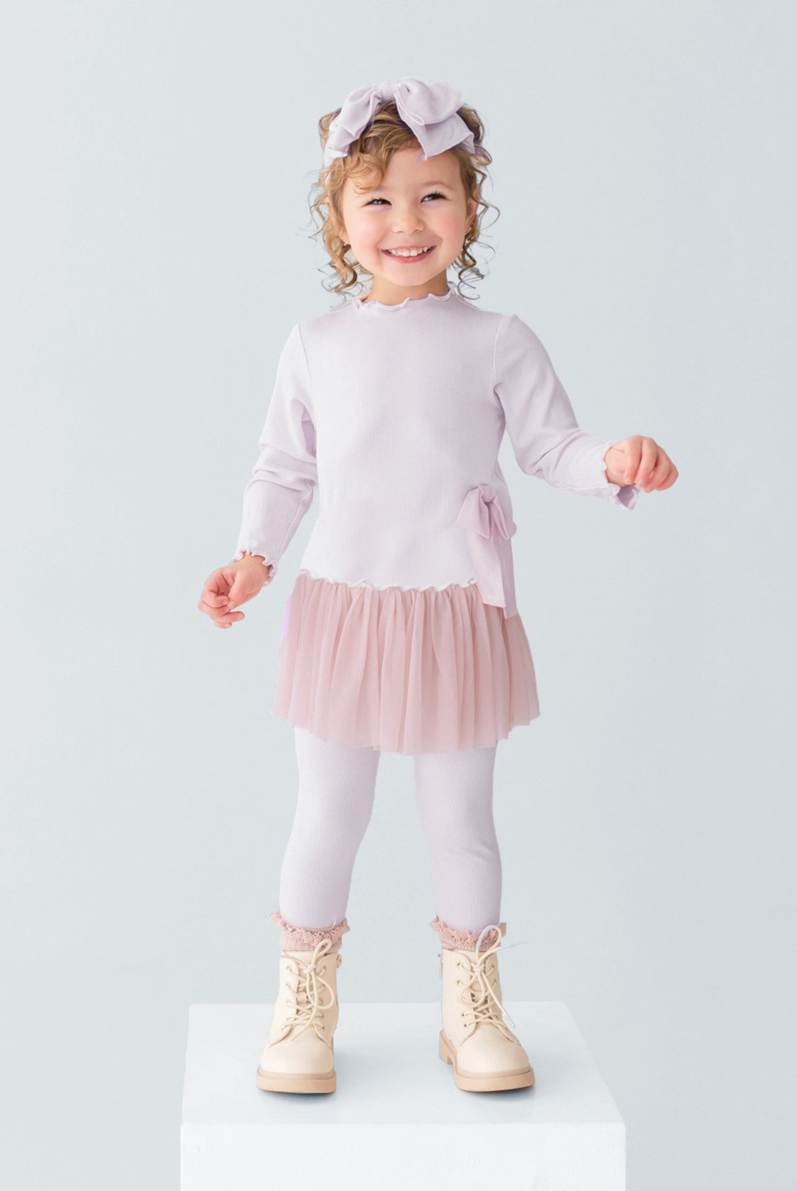 A young girl smiles on a white platform, wearing the Isobella and Chloe Ballerina Two Piece Pant Set with a tulle skirt, ribbed leggings, cream boots, and a large bow headband. The background is plain light gray.