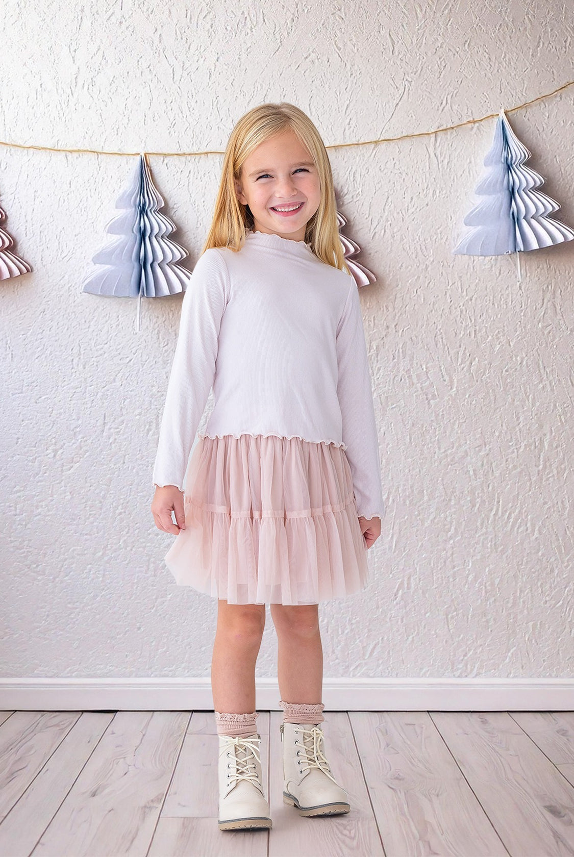 A young girl beams indoors in the Isobella and Chloe Ballerina Two Piece Set—featuring a white long-sleeve top and light pink tulle skirt—paired with white boots and lace socks, with paper tree decorations hanging behind her.