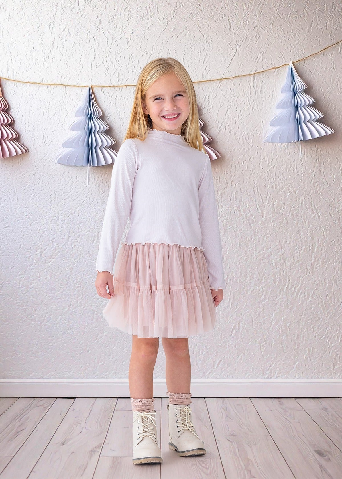 A young girl beams indoors in the Isobella and Chloe Ballerina Two Piece Set—featuring a white long-sleeve top and light pink tulle skirt—paired with white boots and lace socks, with paper tree decorations hanging behind her.
