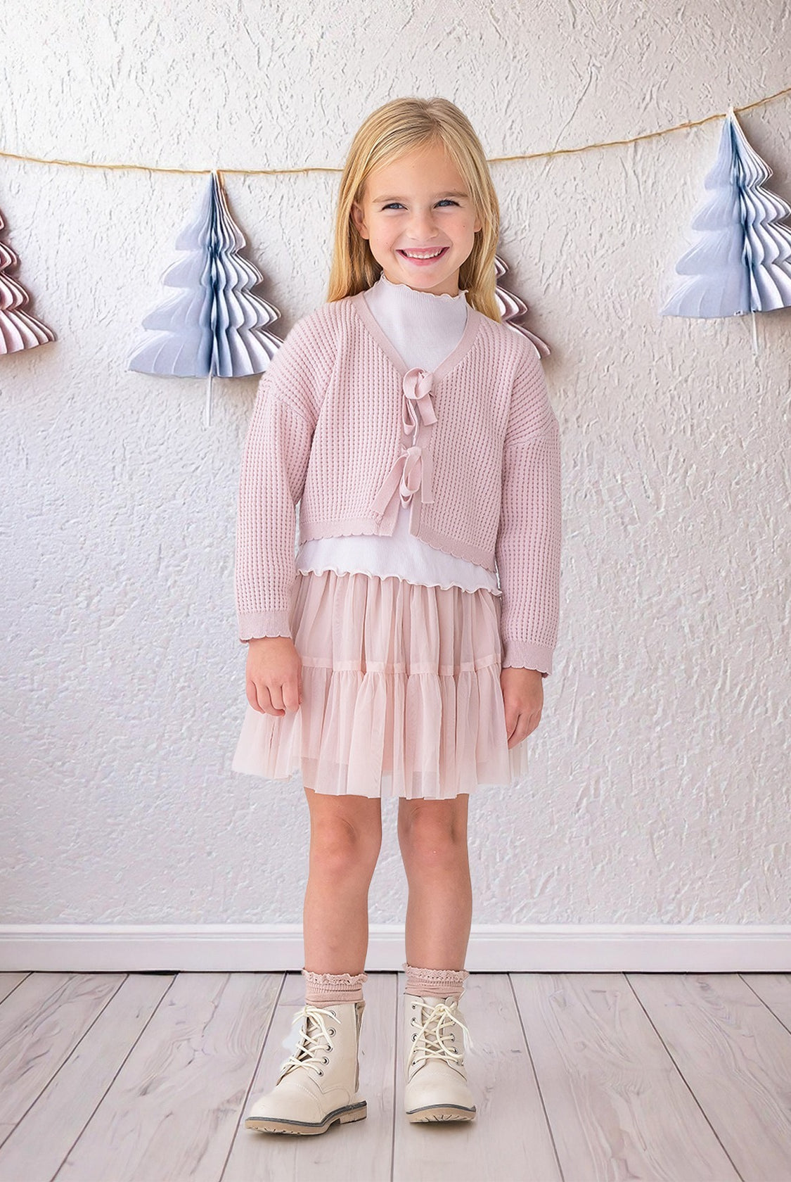 A young girl with blonde hair smiles indoors, wearing the Isobella and Chloe Ballerina Sweater over a white top, paired with a pale pink tulle skirt, sparkly socks, and white boots. A wall with paper Christmas tree garlands is behind her.