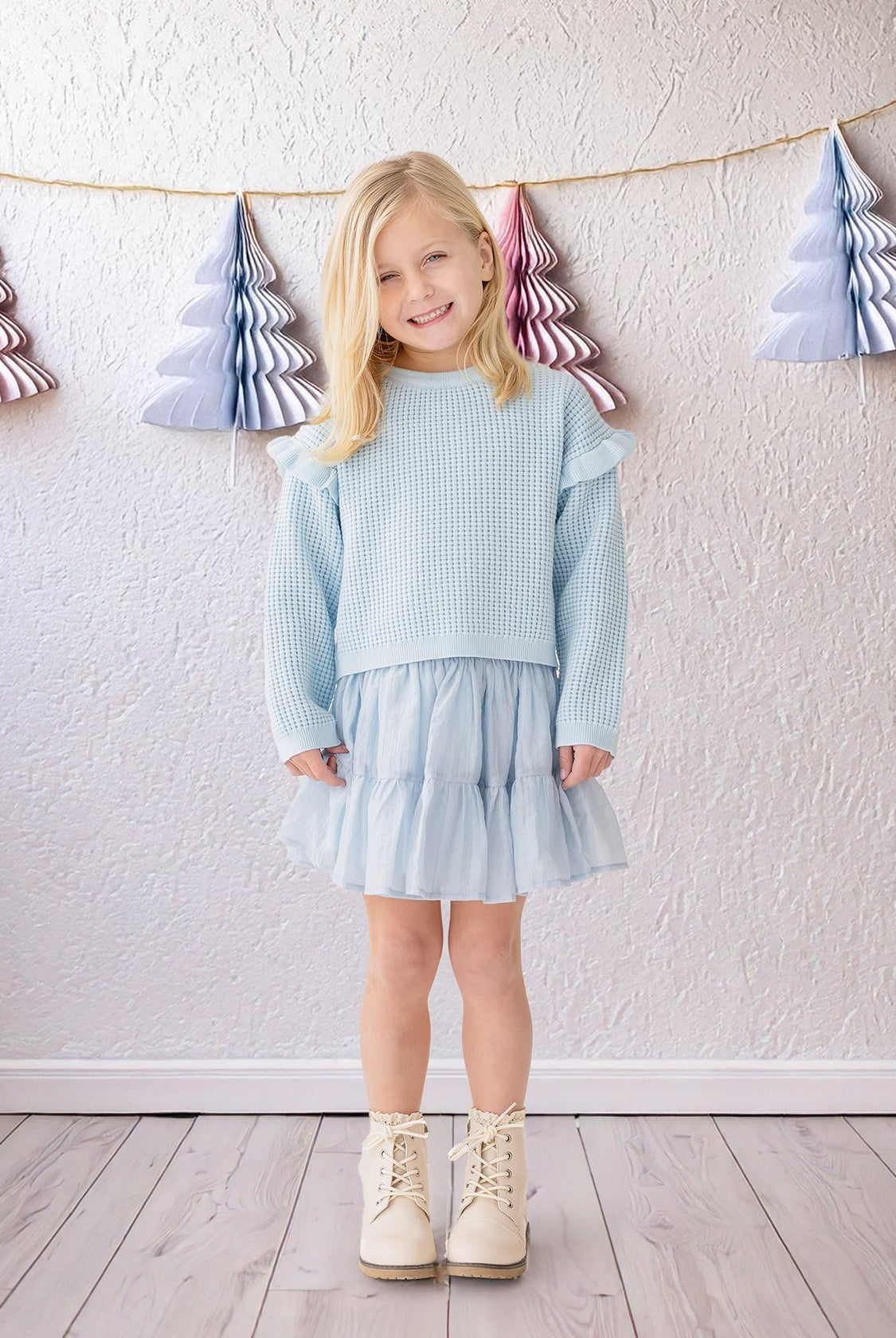 A young blonde girl wears the Isobella and Chloe "Tutu in the Clouds" two-piece set indoors on a wooden floor, with pastel paper Christmas trees hanging on strings against a textured white wall in the background.