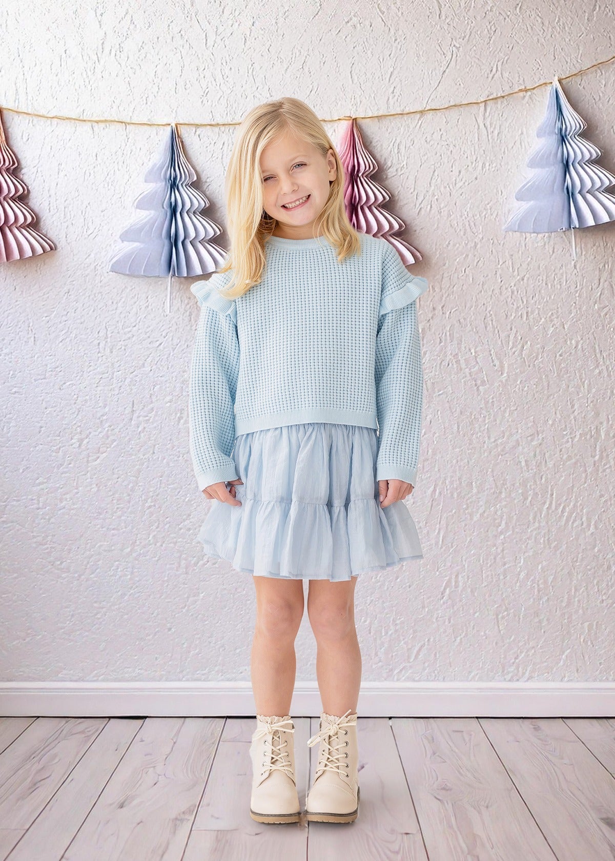 A young blonde girl wears the Isobella and Chloe "Tutu in the Clouds" two-piece set indoors on a wooden floor, with pastel paper Christmas trees hanging on strings against a textured white wall in the background.