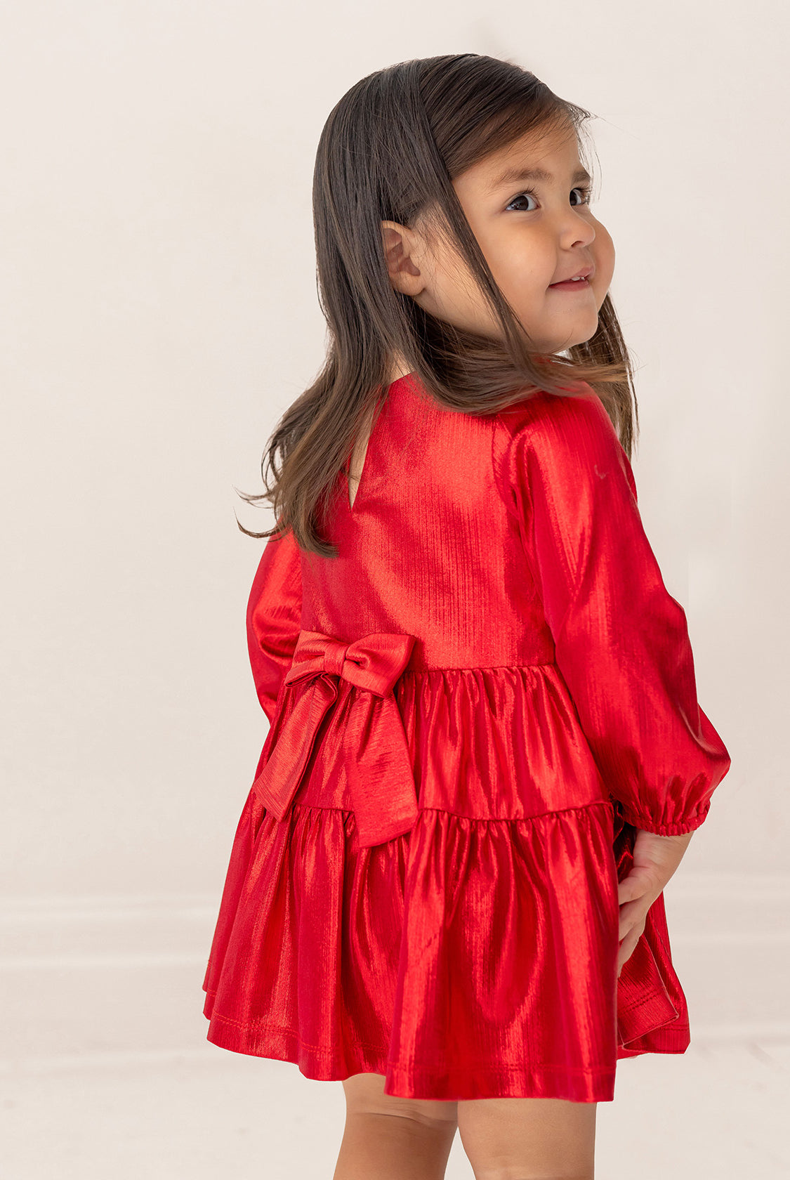 A young girl in the Isobella and Chloe Ruby Red Tiered Dress, featuring a large back bow, stands facing away with a smile over her shoulder. She has long dark hair, and the background is plain and light-colored.