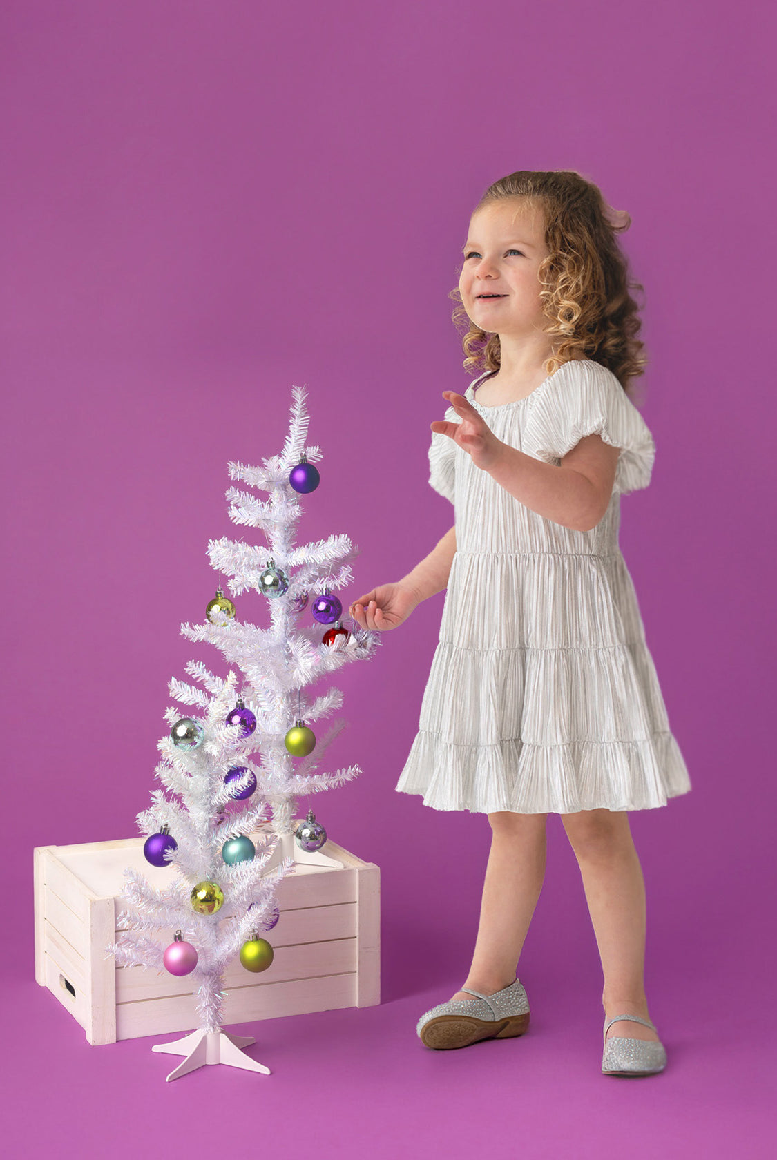 A young child wears the Sparkle and Spin Silver Dress by Isobella and Chloe, standing beside a small white Christmas tree with colorful ornaments, set on a wooden crate against a purple background.
