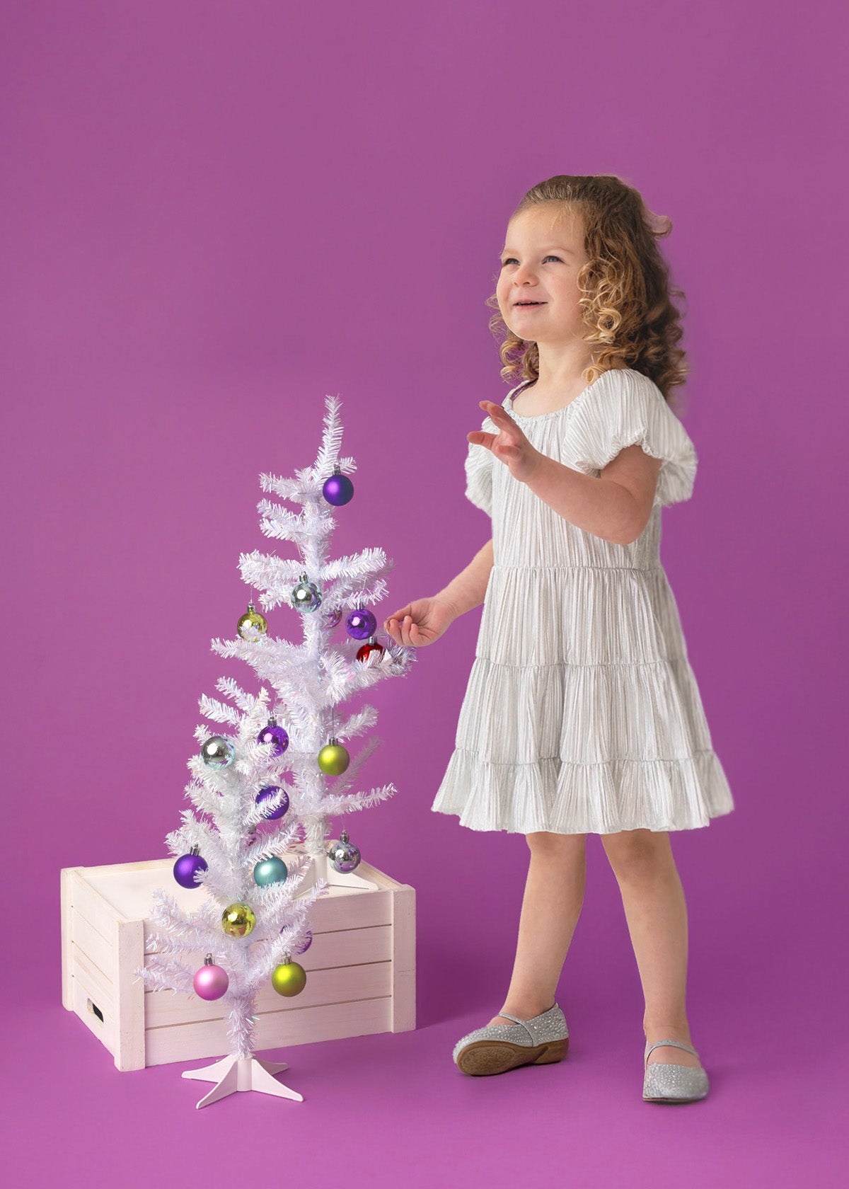 A young child wears the Sparkle and Spin Silver Dress by Isobella and Chloe, standing beside a small white Christmas tree with colorful ornaments, set on a wooden crate against a purple background.