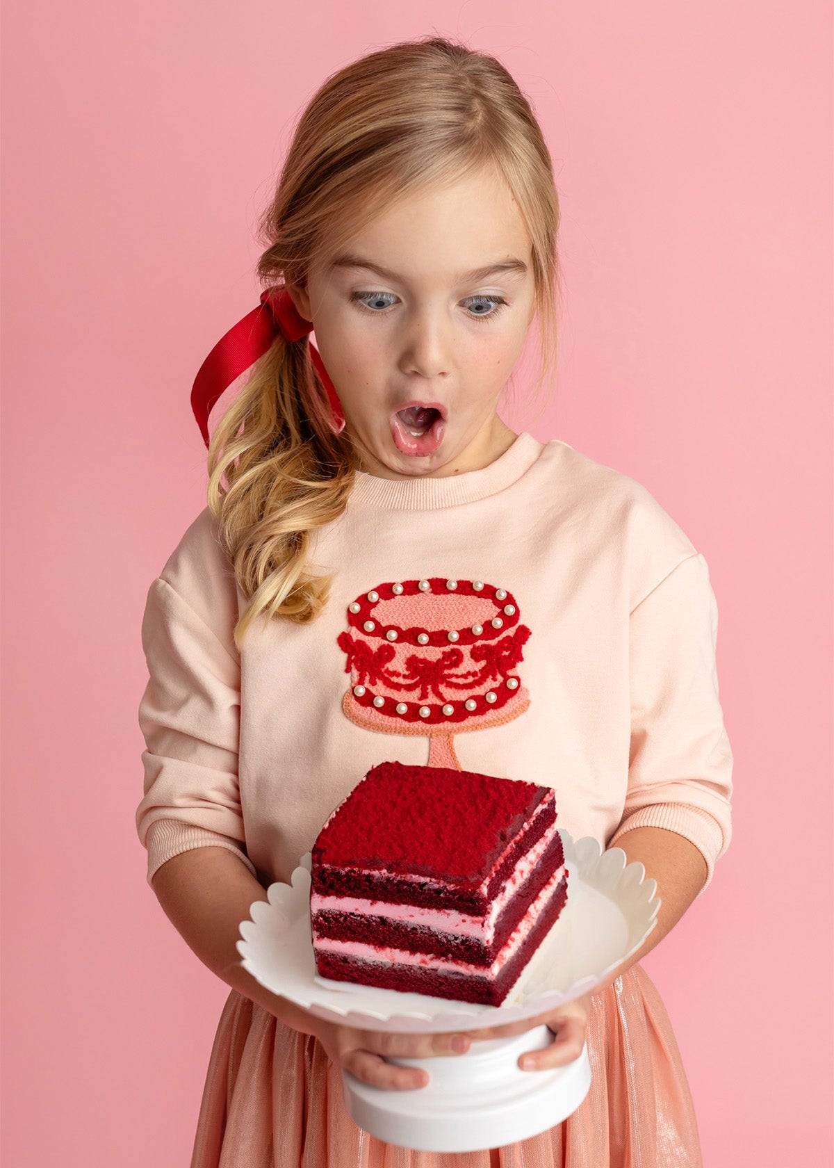A young blond girl with a red ribbon, wearing the Isobella and Chloe Sweet Cakes Two Piece Set, looks surprised and excited as she holds a plate with red velvet cake against a pink background.