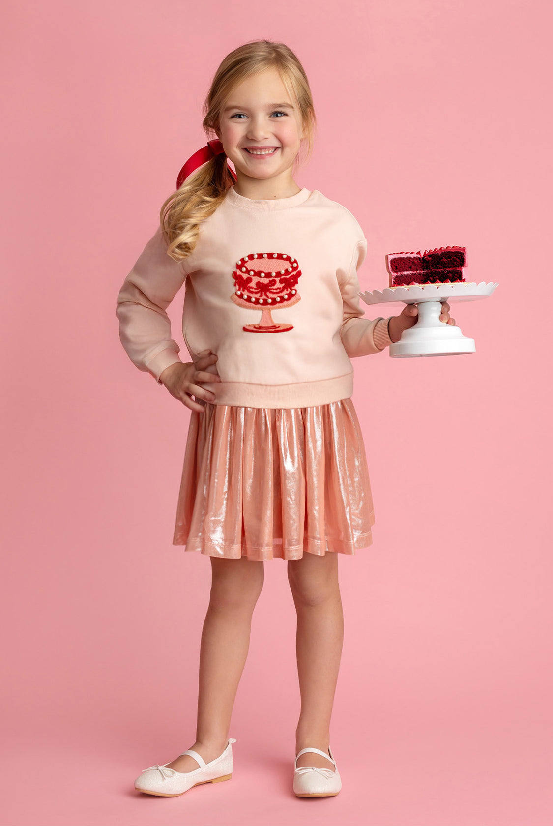 A smiling young girl stands against a pink background, wearing the Isobella and Chloe Sweet Cakes Two Piece Set, featuring an embroidered cake sweater and shiny pink skirt, while holding a small cake with red decoration on a white stand.