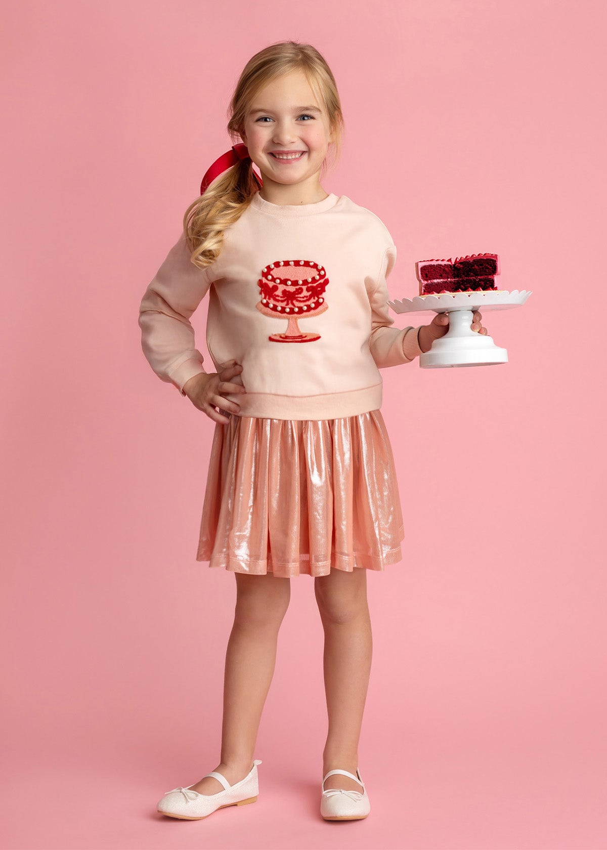 A smiling young girl stands against a pink background, wearing the Isobella and Chloe Sweet Cakes Two Piece Set, featuring an embroidered cake sweater and shiny pink skirt, while holding a small cake with red decoration on a white stand.