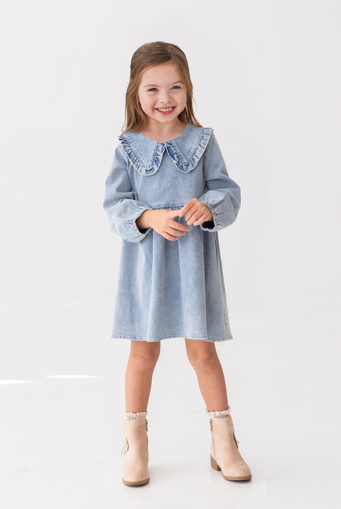 A young girl with light brown hair smiles while standing against a white background, wearing the Mabel and Honey Denim Days Long Sleeve Dress styled with beige ankle boots.