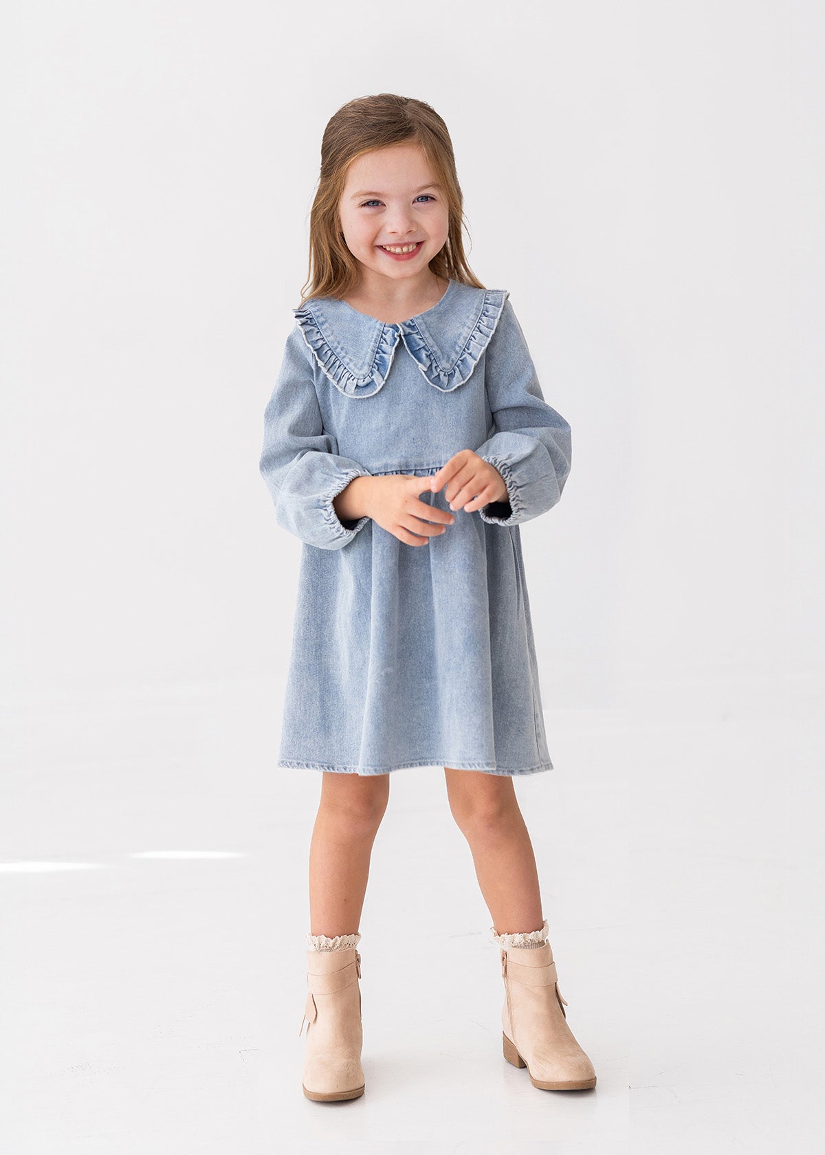 A young girl with light brown hair smiles while standing against a white background, wearing the Mabel and Honey Denim Days Long Sleeve Dress styled with beige ankle boots.