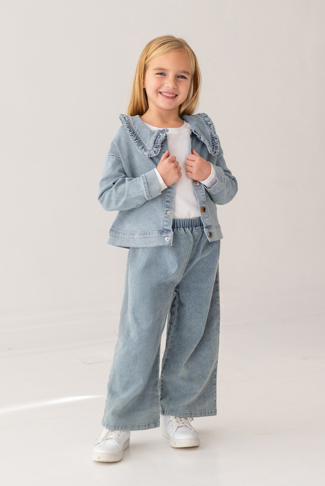 A young girl with blonde hair smiles in the Mabel and Honey Denim Days Ruffled Collar Jacket, paired with matching wide-leg denim pants, a white shirt, and white sneakers against a plain light background.