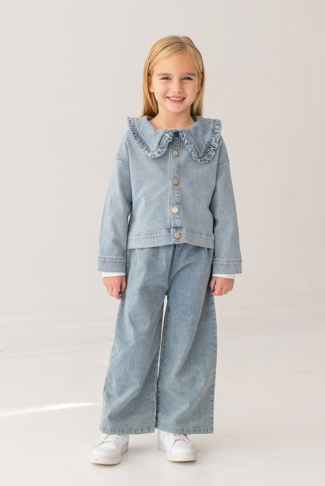 A young blonde girl smiles in a Mabel and Honey Denim Days Ruffled Collar Jacket, paired with matching wide-leg denim pants and white sneakers, standing against a plain light background—a stylish kids’ wardrobe choice.