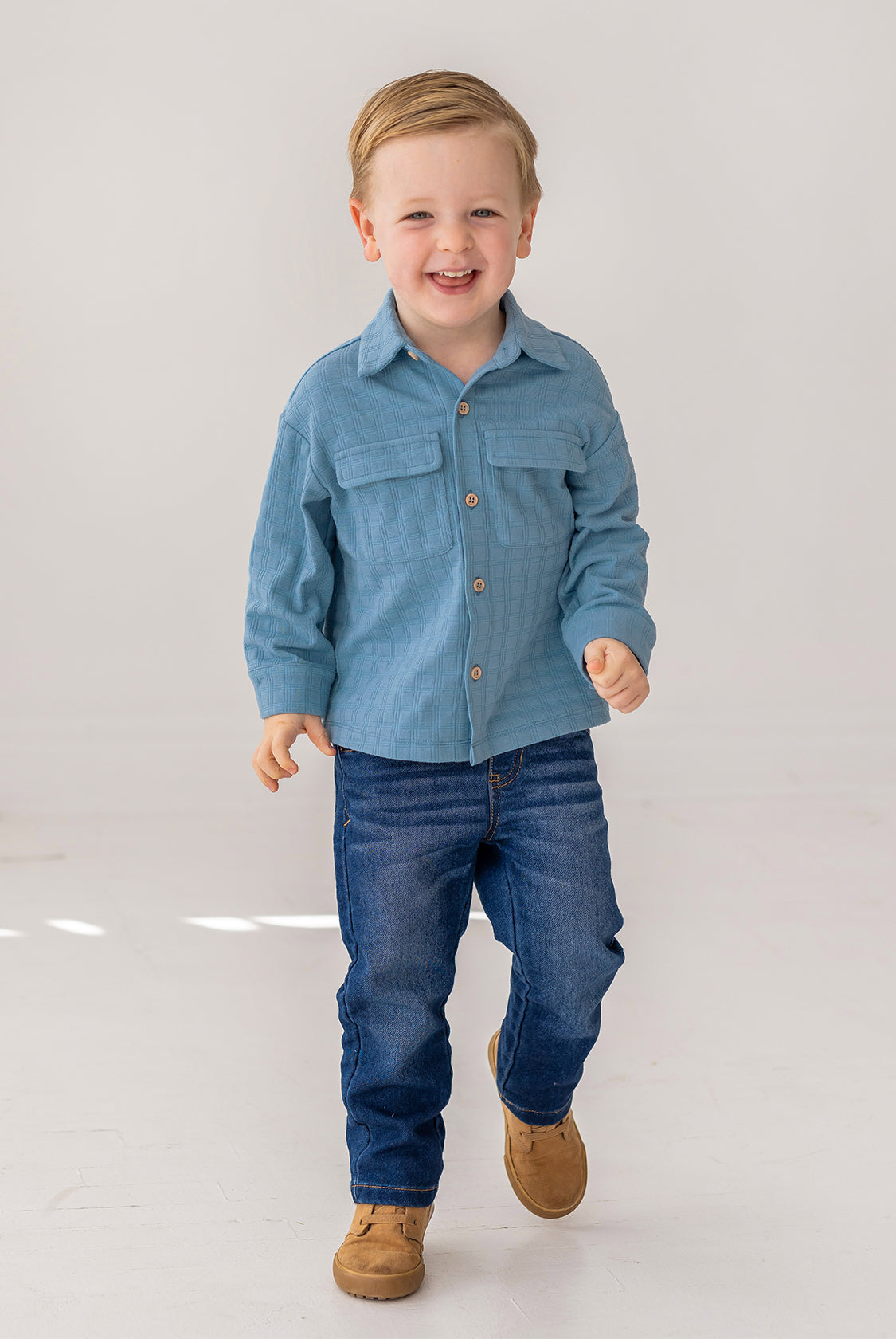A young boy with short light brown hair smiles as he walks, wearing the Beckett and Bear Lucas Jacket over a light blue shirt, dark blue jeans, and brown shoes against a plain white background.