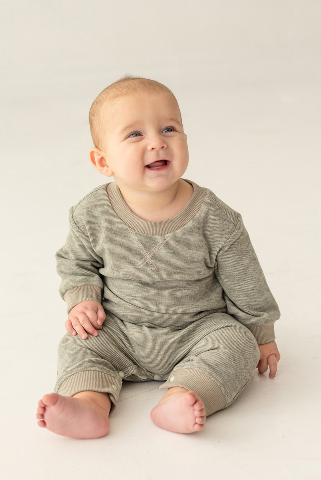 A smiling baby with light skin and short hair sits on a light-colored floor, wearing the Miles Romper by Beckett and Bear—a sage green, minimalist-inspired boys romper with ribbed fabric and matching pants.
