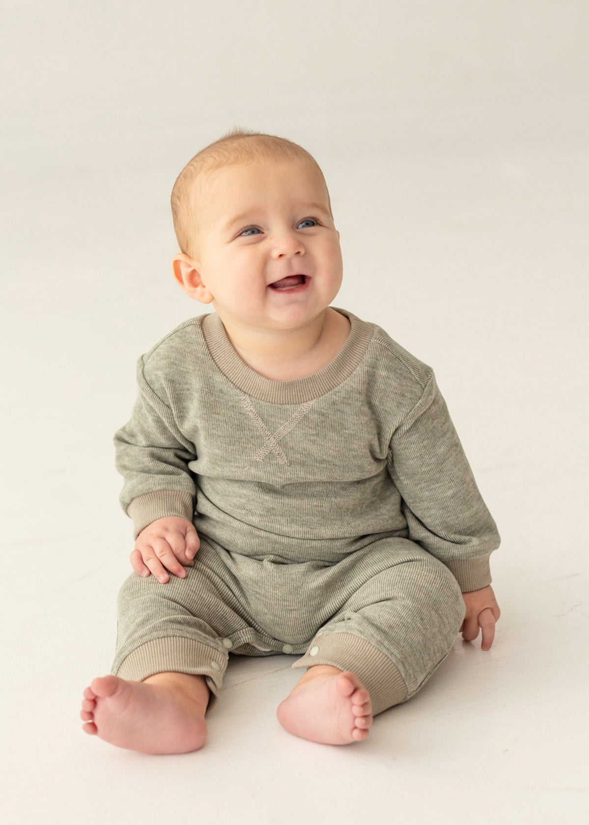 A smiling baby with light skin and short hair sits on a light-colored floor, wearing the Miles Romper by Beckett and Bear—a sage green, minimalist-inspired boys romper with ribbed fabric and matching pants.