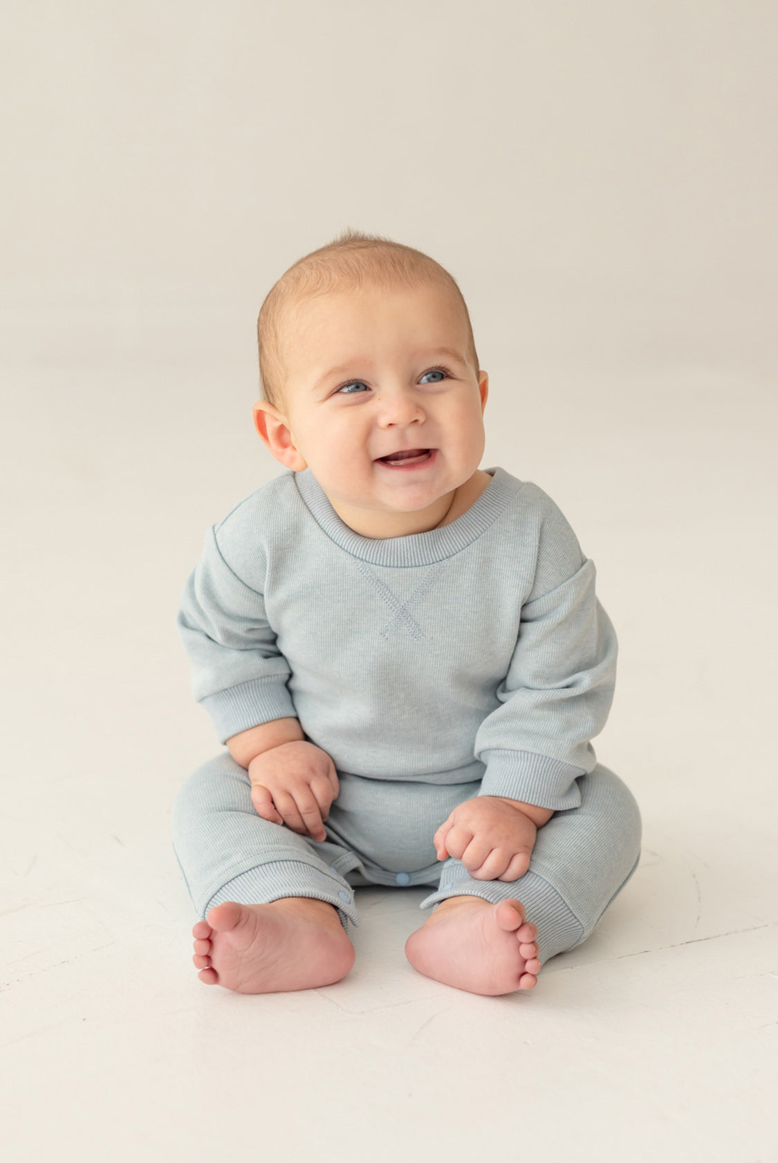 A baby with light hair and blue eyes smiles and looks to the side, sitting on the floor in the Beckett and Bear Aiden Romper against a simple, light background.