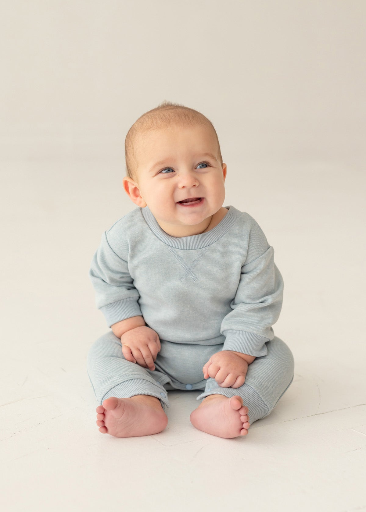 A baby with light hair and blue eyes smiles and looks to the side, sitting on the floor in the Beckett and Bear Aiden Romper against a simple, light background.