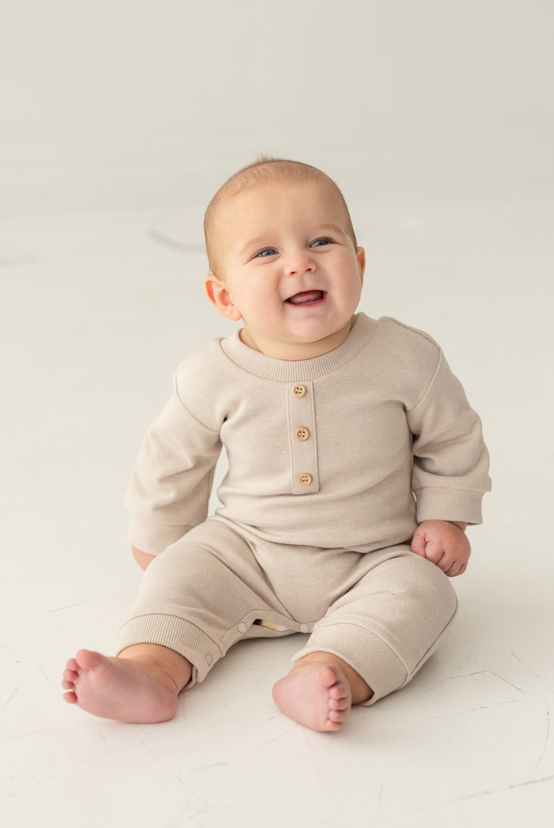 A smiling baby sits barefoot on a light floor against a neutral background, wearing the neutral-tone Isaac Romper by Beckett and Bear, featuring wooden buttons.