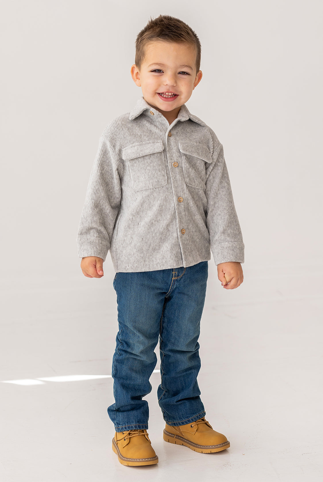 A young boy with short brown hair smiles while standing in a light gray Beckett and Bear Grayson Jacket over a button-up shirt, blue jeans, and tan shoes, set against a plain white background.