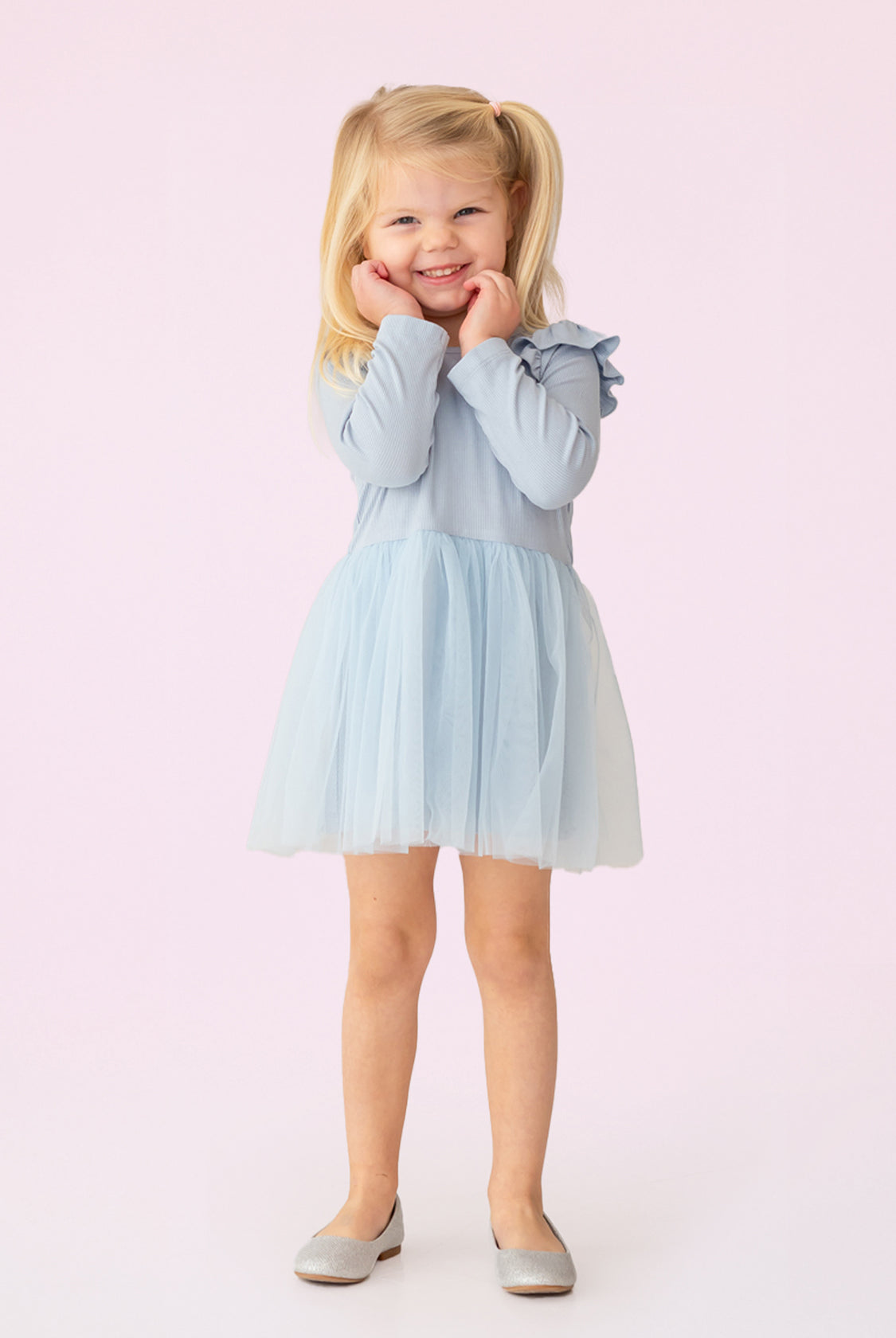 A young girl with blonde pigtails smiles against a pale pink background, wearing the Isobella and Chloe Tutu in the Clouds Dress, with its light blue tulle skirt and long-sleeved ribbed bodice, her hands gently touching her cheeks.