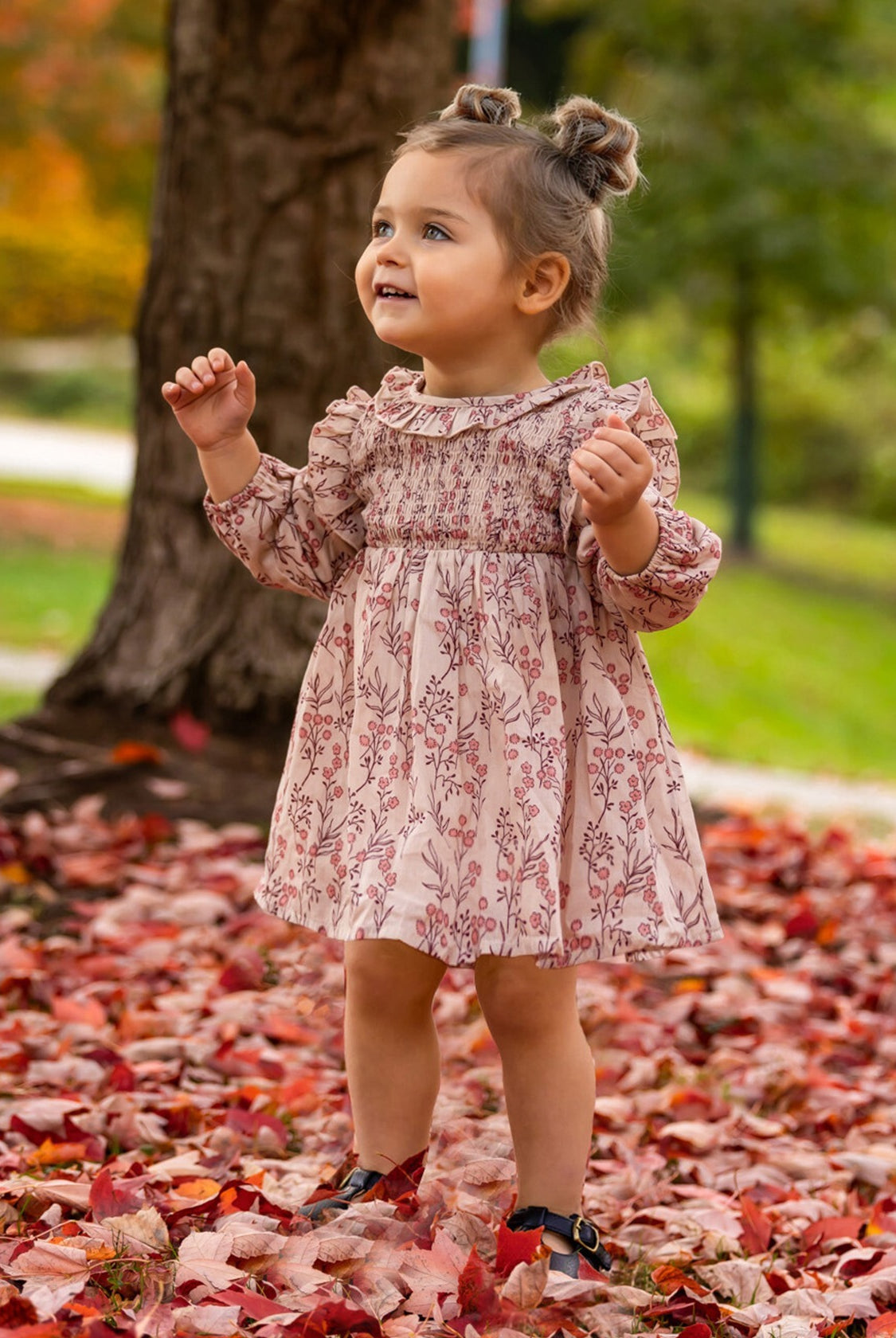 A young girl with brown hair in two buns smiles outdoors among autumn leaves, wearing the Mabel + Honey Autumn Floral Tan Long Sleeve Dress and black shoes. A tree trunk and green foliage are visible in the background.