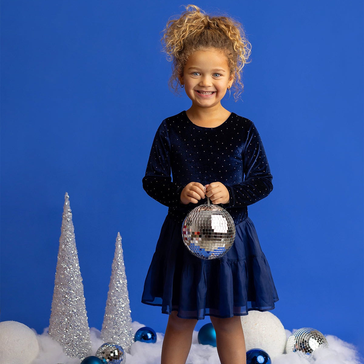 A young girl wearing the Isobella and Chloe Velvet Dreams Long Sleeve Rhinestone Studded Tiered Skirt Dress holds a disco ball, standing in front of silver decorations, blue ornaments, and faux snow, all set against a blue background.