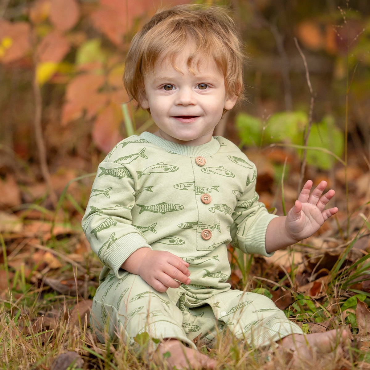 A smiling toddler with light brown hair sits on the grass, wearing the Beckett and Bear Matthew Long Sleeve Fish Print Ribbed Trim Button Placket Romper. Autumn leaves and plants are softly blurred in the background as the child waves with one hand.