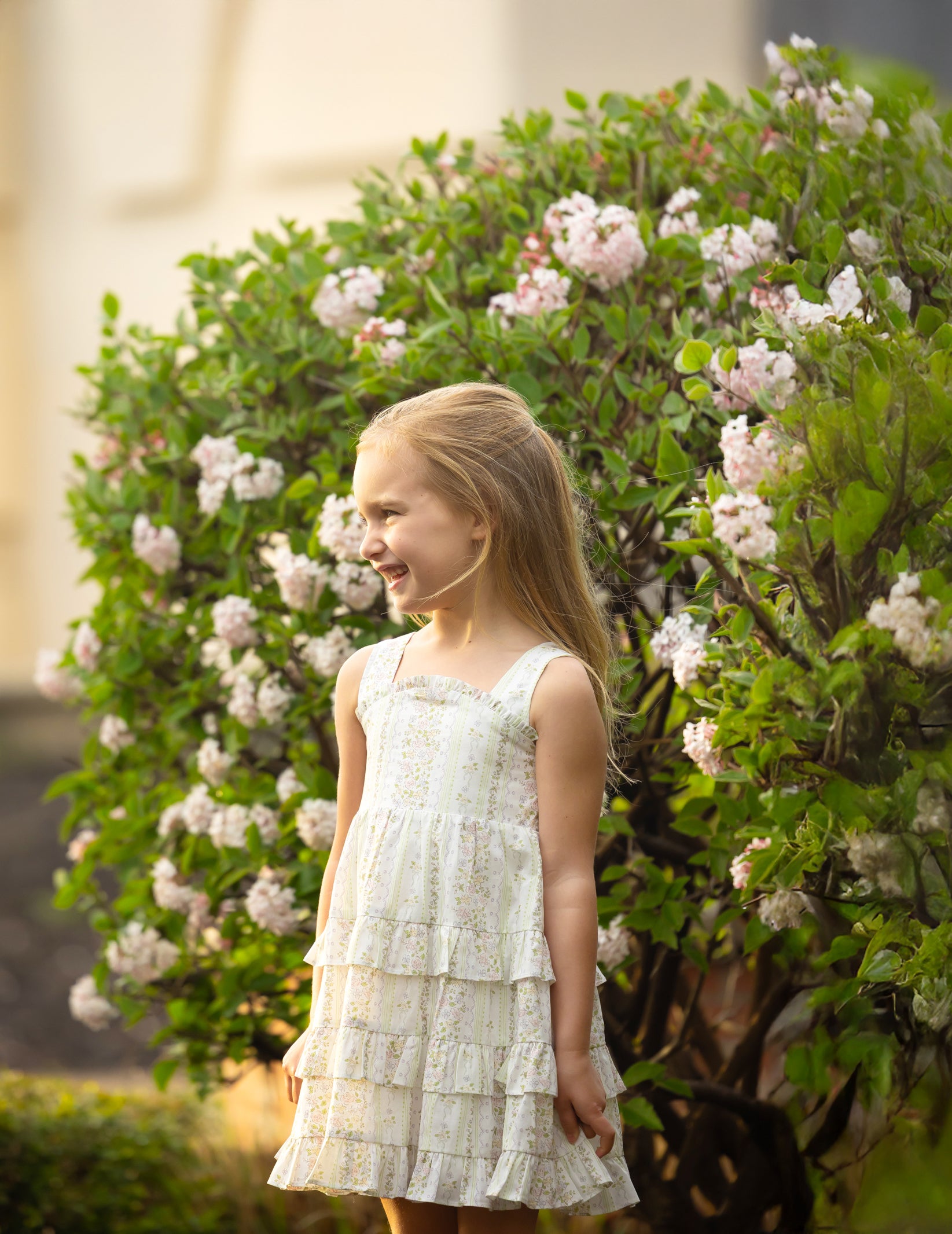 A young girl in a tiered floral dress smiling in front of a blooming flowering bush.