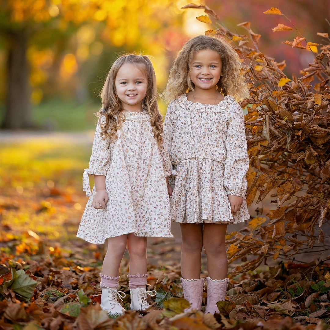 Two young girls in matching floral dresses standing in a park covered in autumn leaves.