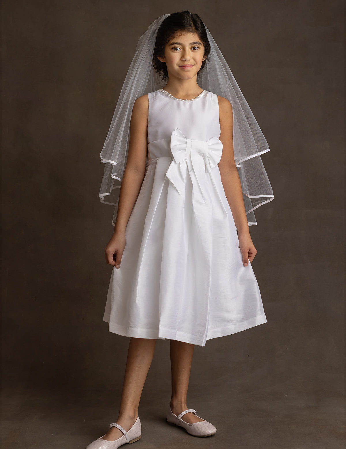 A young girl wears the Mabel and Honey Taffeta Bow Communion Dress with a beaded pearl neckline, large front bow, white veil, and light flats, standing against a dark background.