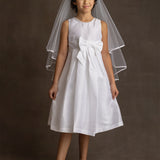 A young girl wears the Mabel and Honey Taffeta Bow Communion Dress with a beaded pearl neckline, large front bow, white veil, and light flats, standing against a dark background.
