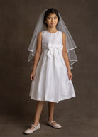 A young girl wears the Mabel and Honey Taffeta Bow Communion Dress with a beaded pearl neckline, large front bow, white veil, and light flats, standing against a dark background.