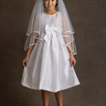 A young girl smiles softly at the camera in a Mabel and Honey Taffeta Bow Communion Dress with a beaded pearl neckline, a large waist bow, white veil, and light pink ballet flats against a plain brown background.