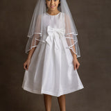 A young girl smiles softly at the camera in a Mabel and Honey Taffeta Bow Communion Dress with a beaded pearl neckline, a large waist bow, white veil, and light pink ballet flats against a plain brown background.
