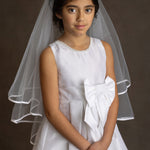 A young girl wears the Mabel and Honey Taffeta Bow Communion Dress with a beaded pearl neckline and sheer white veil, standing against a dark background with her hands folded.