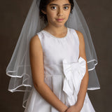 A young girl wears the Mabel and Honey Taffeta Bow Communion Dress with a beaded pearl neckline and sheer white veil, standing against a dark background with her hands folded.