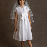 A young girl with dark hair smiles in the Mabel and Honey Taffeta Bow Communion Dress with beaded pearl neckline, a short white bolero, veil, and light flats, posing against a plain dark background.