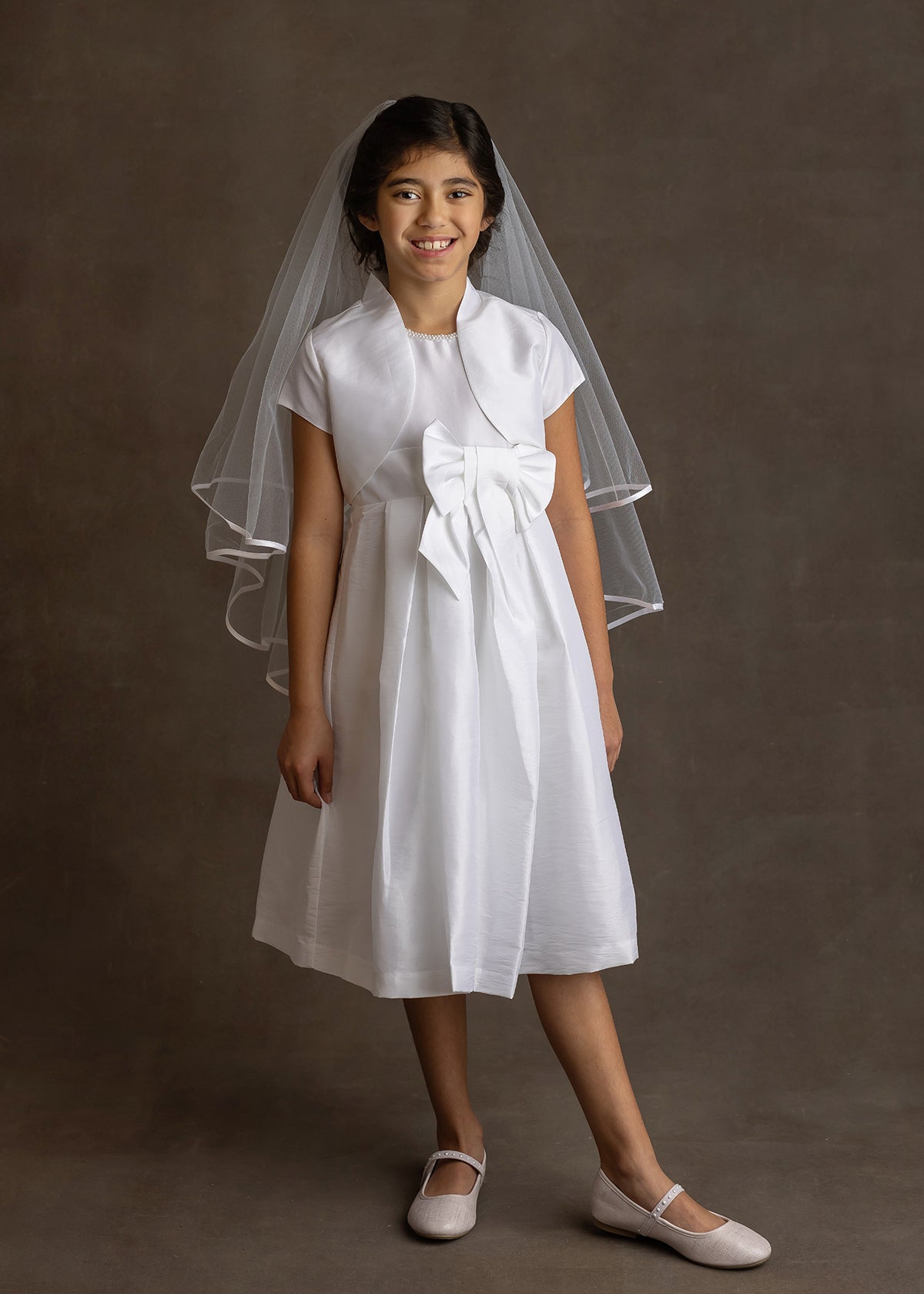 A young girl with dark hair smiles in the Mabel and Honey Taffeta Bow Communion Dress with beaded pearl neckline, a short white bolero, veil, and light flats, posing against a plain dark background.