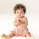 A toddler with curly hair and a pink bow sits on the floor wearing the Mabel and Honey Pink Petals Two Piece Short Short Set and beige shoes, looking slightly upward with hands clasped, against a plain white background.