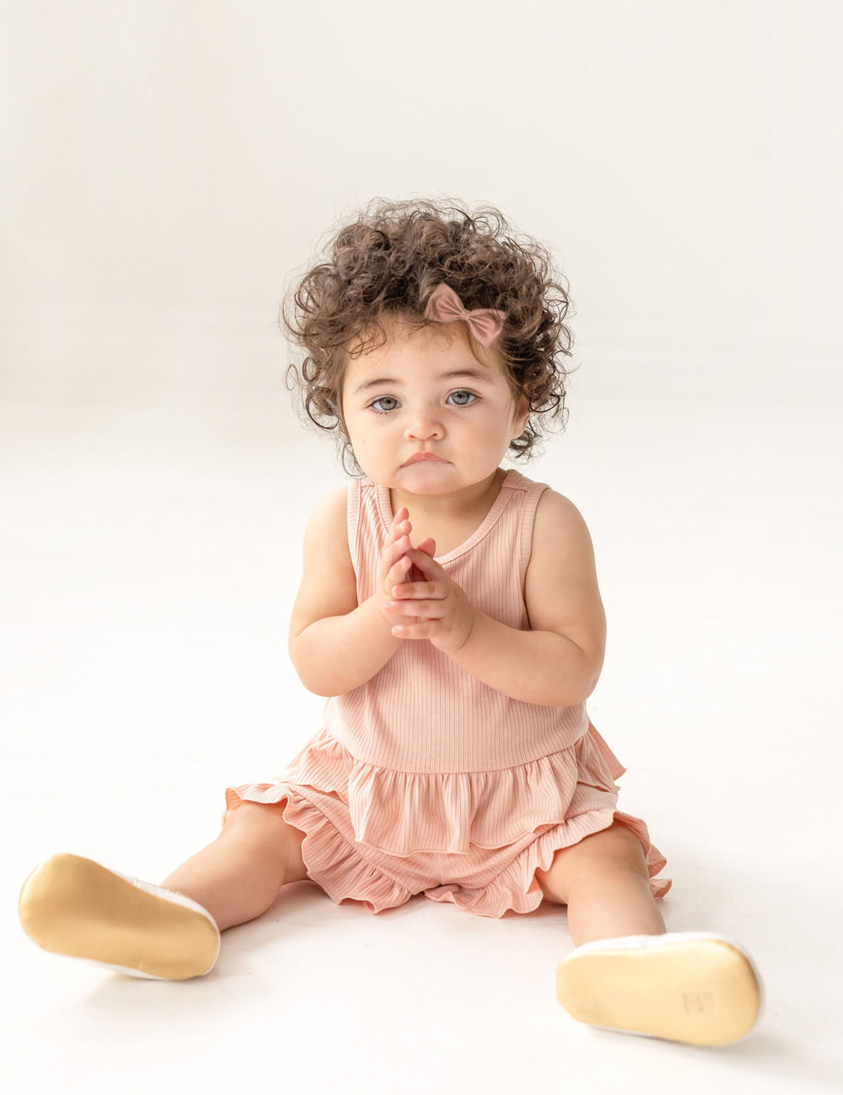 A toddler with curly hair and a pink bow sits on the floor wearing the Mabel and Honey Pink Petals Two Piece Short Short Set and beige shoes, looking slightly upward with hands clasped, against a plain white background.
