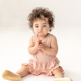 A toddler with curly hair and a pink bow sits on the floor wearing the Mabel and Honey Pink Petals Two Piece Short Short Set and beige shoes, looking slightly upward with hands clasped, against a plain white background.