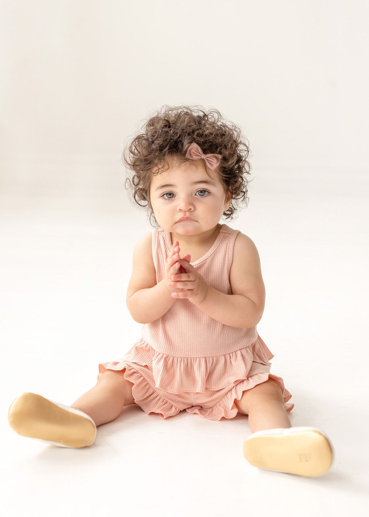 A toddler with curly hair and a pink bow sits on the floor wearing the Mabel and Honey Pink Petals Two Piece Short Short Set and beige shoes, looking slightly upward with hands clasped, against a plain white background.