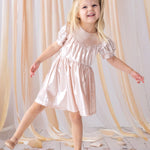 A young girl beams as she poses on one foot in the Isobella and Chloe Golden Glow Pink Dress, featuring shiny metallic pink fabric and puffed short sleeves, with matching shoes. She stands on a white floor amid gold and pink star confetti and peach drapes.