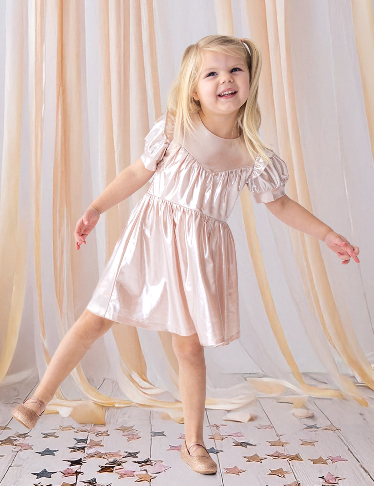 A young girl beams as she poses on one foot in the Isobella and Chloe Golden Glow Pink Dress, featuring shiny metallic pink fabric and puffed short sleeves, with matching shoes. She stands on a white floor amid gold and pink star confetti and peach drapes.