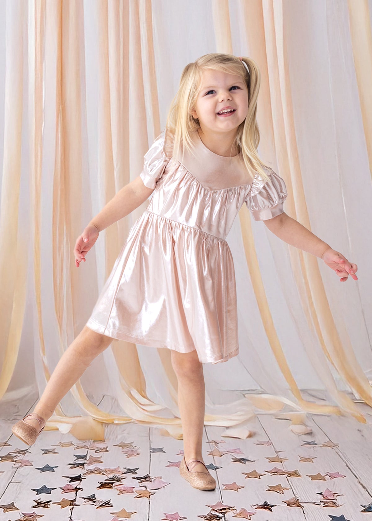 A young girl beams as she poses on one foot in the Isobella and Chloe Golden Glow Pink Dress, featuring shiny metallic pink fabric and puffed short sleeves, with matching shoes. She stands on a white floor amid gold and pink star confetti and peach drapes.