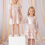 Two young girls hold hands and smile, both wearing Isobella and Chloe’s Golden Glow Pink Dresses with puffed sleeves and matching shoes. One girl stands on a white cube, surrounded by peach ribbons and star confetti for a magical touch.