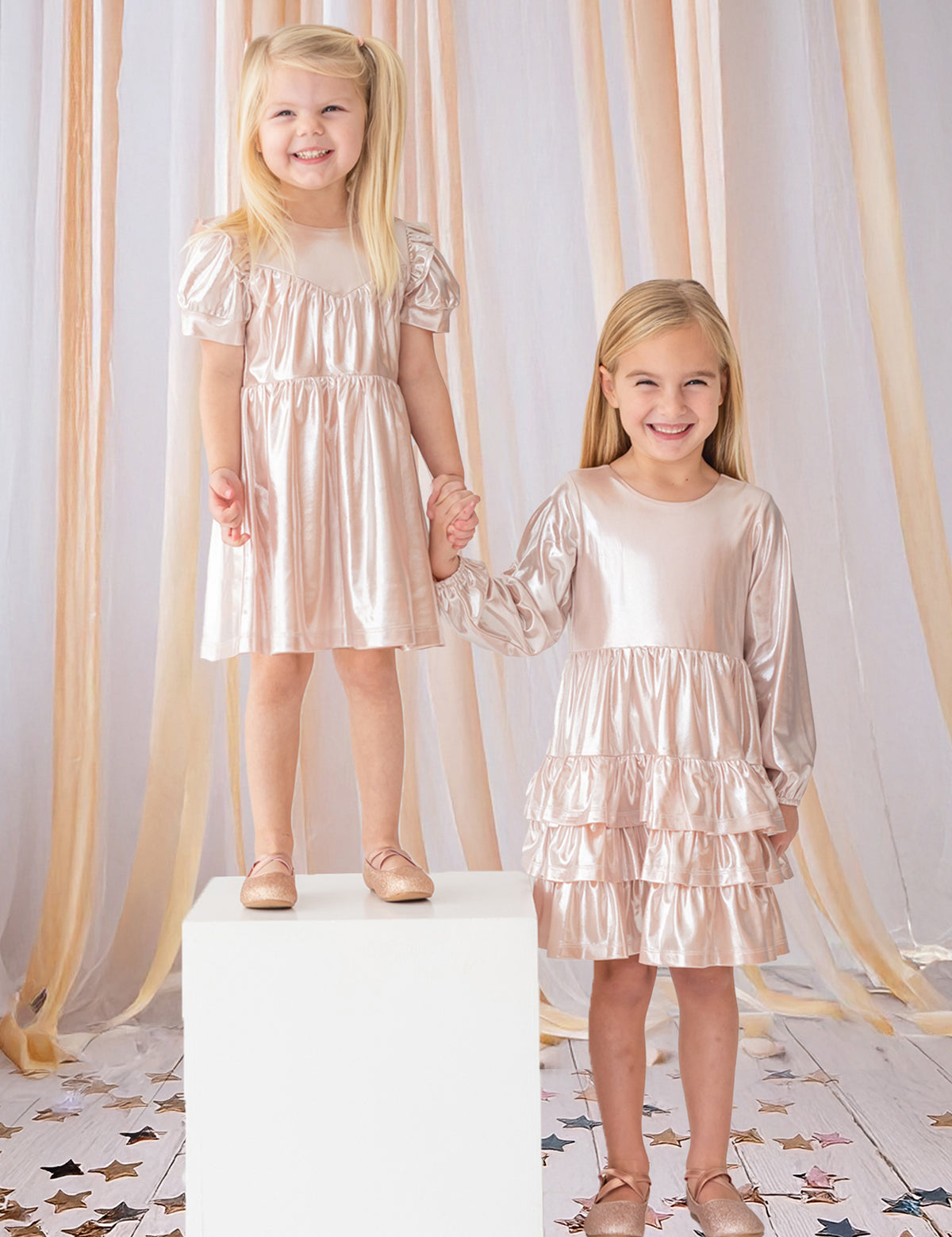 Two young girls hold hands and smile, both wearing Isobella and Chloe’s Golden Glow Pink Dresses with puffed sleeves and matching shoes. One girl stands on a white cube, surrounded by peach ribbons and star confetti for a magical touch.