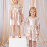 Two young girls hold hands and smile, both wearing Isobella and Chloe’s Golden Glow Pink Dresses with puffed sleeves and matching shoes. One girl stands on a white cube, surrounded by peach ribbons and star confetti for a magical touch.